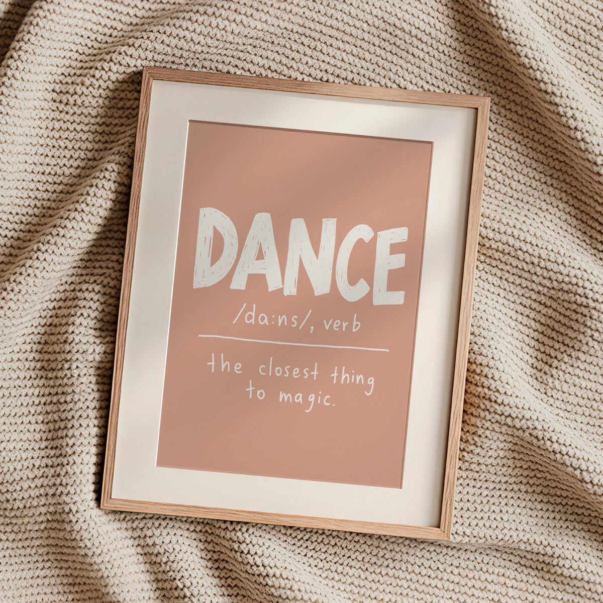 A framed typographic art print reading “DANCE /dæns/, verb” and “the closest thing to magic.” The print has a dusty pink background with white lettering, set in a light wood frame on a textured beige knit blanket.