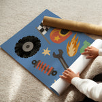 A person unrolling a large blue wall mural or poster featuring automotive-themed illustrations like a tire, wrench, flame, gear, and checkered flag, on a beige carpet. A brown cardboard roll is partially visible.