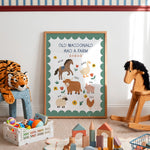 A framed children's educational poster reading 'OLD MACDONALD HAD A FARM' with illustrated farm animals and flowers, displayed in a styled nursery or playroom setting with toys around it.