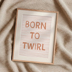 A framed typographic wall art print reading “BORN TO TWIRL” in peach lettering over a pale striped background, set in a light wood frame on a textured cream knit blanket.