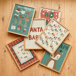 Six framed Christmas-themed art prints arranged in a flat-lay on a light wood floor. The prints feature festive illustrations including Santa, a reindeer, holiday letters, and seasonal phrases, each in a light natural wood frame.