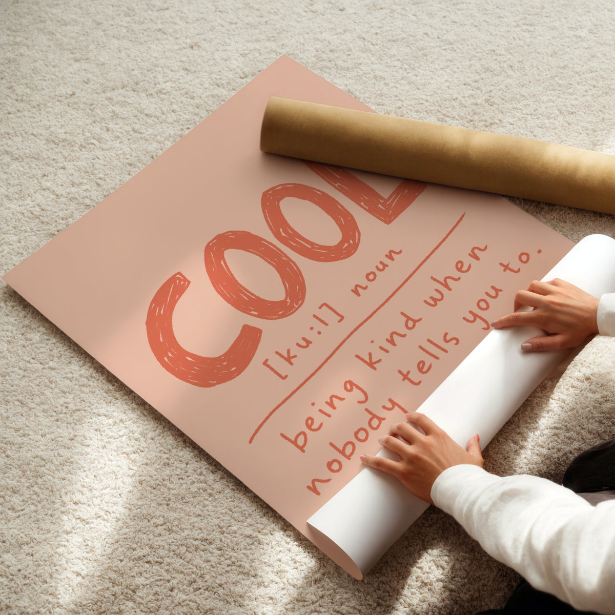 Hands unrolling cool definition print from kraft shipping tube, showing terracotta lettering on blush