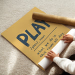 A person unrolling a large yellow wall art poster on a beige carpet, with a brown cardboard mailing tube placed on top. The poster features the word “PLAY” in large dark blue letters with a handwritten-style definition underneath.