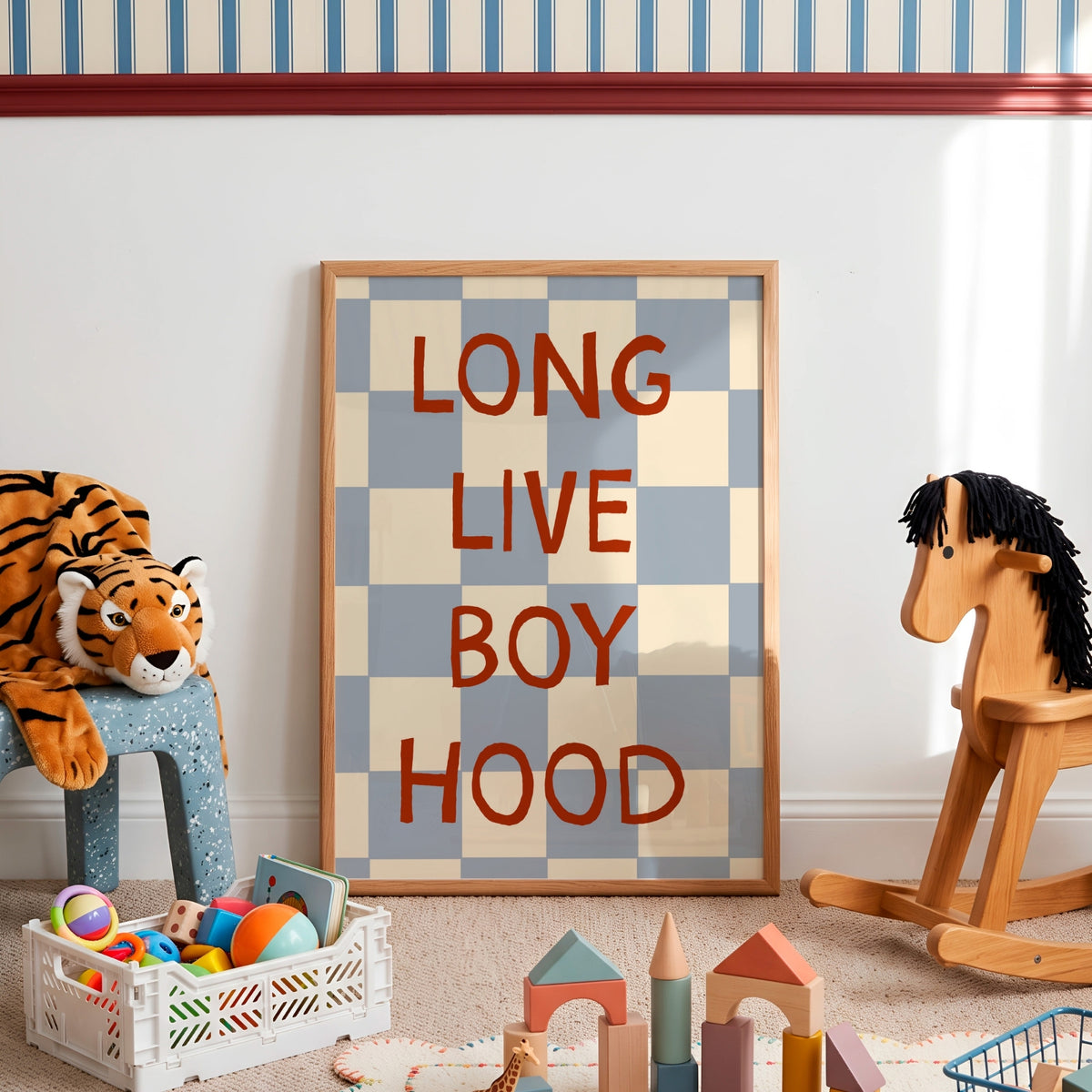 A framed poster with the text 'LONG LIVE BOY HOOD' in red letters over a blue and beige checkered pattern, leaning against a white wall in a styled children's room with toys and a rocking horse.