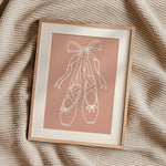 A framed minimalist art print featuring a white line drawing of a ballerina's tutu on a dusty pink background, styled on a textured beige knit blanket.