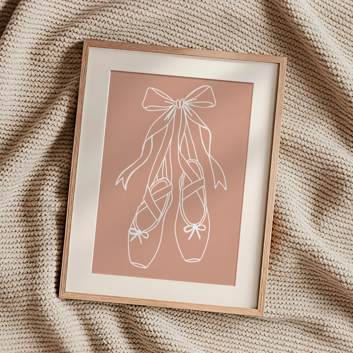 A framed minimalist art print featuring a white line drawing of a ballerina's tutu on a dusty pink background, styled on a textured beige knit blanket.