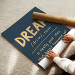A navy blue wall art poster with gold lettering reading 'DREAM a really good idea that hasn't happened yet' is being unrolled on a beige carpet. A person's hands and arms are visible pulling the white backing paper, and a brown cardboard mailing tube lies nearby.