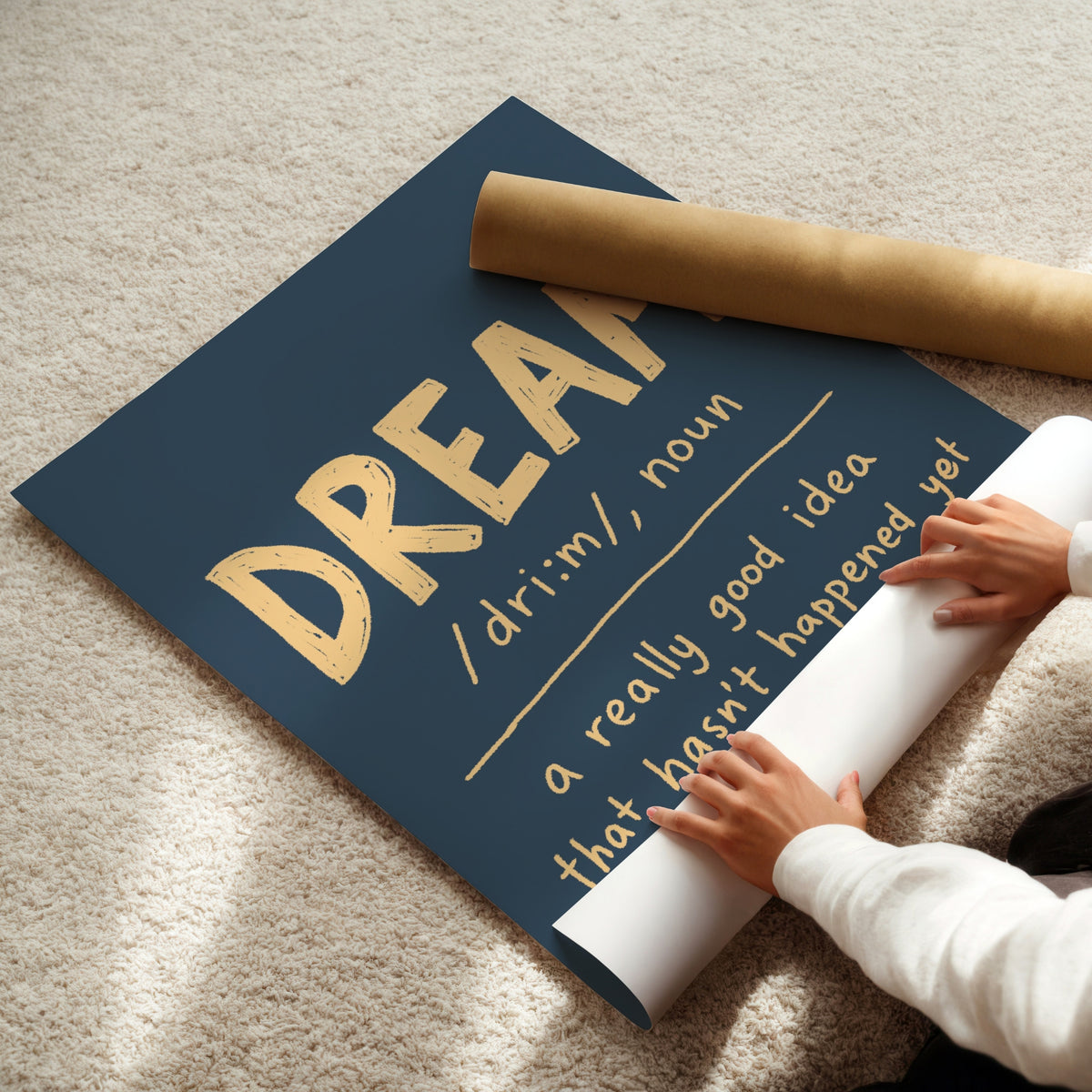 A navy blue wall art poster with gold lettering reading 'DREAM a really good idea that hasn't happened yet' is being unrolled on a beige carpet. A person's hands and arms are visible pulling the white backing paper, and a brown cardboard mailing tube lies nearby.