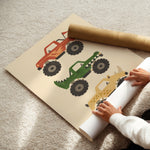 A large printed poster or play mat featuring three colorful monster truck illustrations, laid on a beige carpet with a person's hands unrolling the bottom edge. A brown cardboard mailing tube rests across the top.