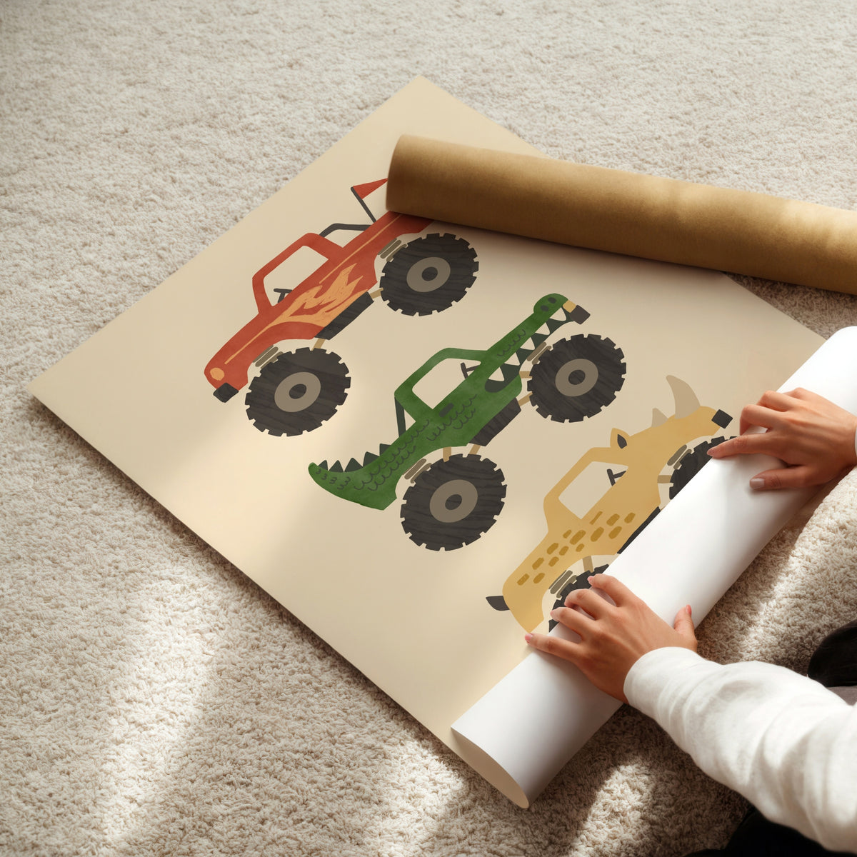A large printed poster or play mat featuring three colorful monster truck illustrations, laid on a beige carpet with a person's hands unrolling the bottom edge. A brown cardboard mailing tube rests across the top.