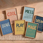 Six framed typographic wall art prints arranged on a woven beige rug over a wood floor. The prints feature words such as COOL, MESS, BRAVE, PLAY, MAGIC, and DREAM with handwritten-style definitions in contrasting colors inside light wood frames.