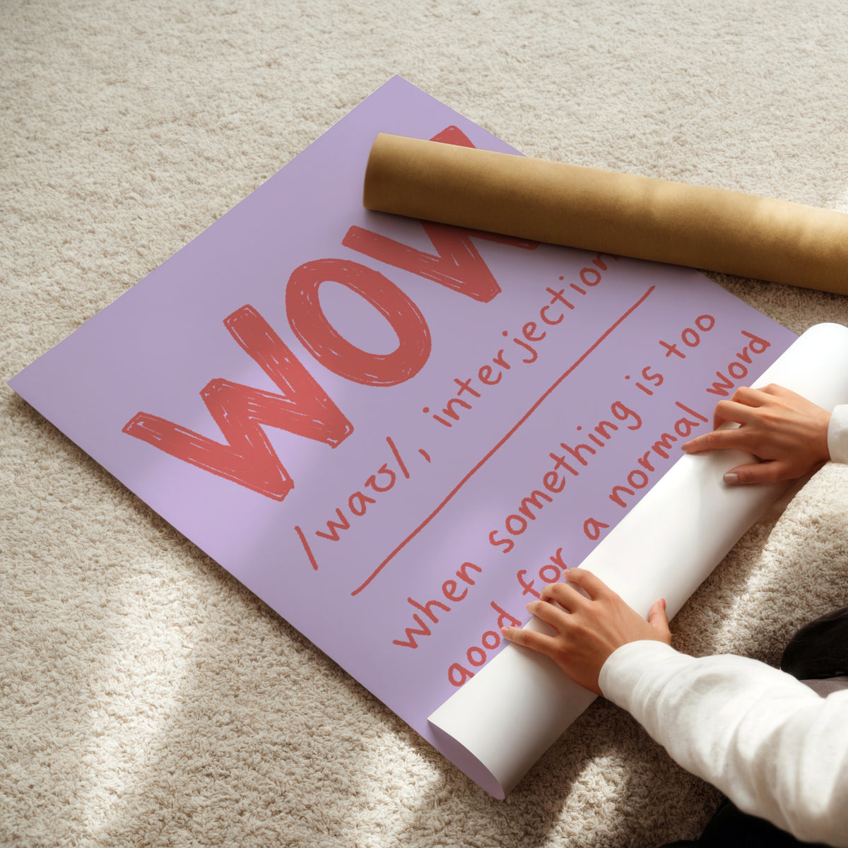 A person unrolls a large lavender poster on a beige carpet, revealing red handwritten-style text that reads 'WOW /wɒ/ when something is too good for a normal word.' A brown cardboard mailing tube rests across the poster.