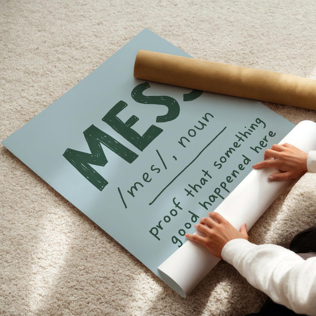 A person unrolling a large typographic wall art print on a beige carpet, with a brown cardboard mailing tube placed on top. The print features the word “MESS” in large dark green letters with a dictionary-style definition underneath.