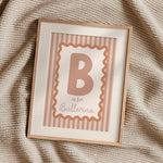 A framed personalized baby nursery print featuring the letter 'B' and the text 'is for Ballerina' in a dusty rose color, set in a light wood frame on a textured beige knit blanket.