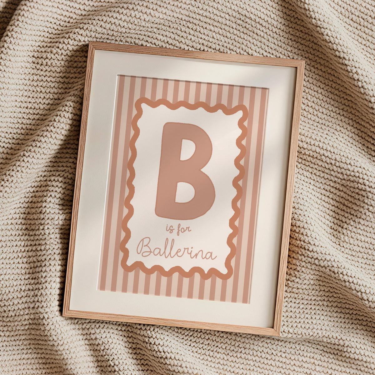 A framed personalized baby nursery print featuring the letter 'B' and the text 'is for Ballerina' in a dusty rose color, set in a light wood frame on a textured beige knit blanket.