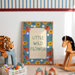 A framed children's art print reading 'LITTLE WILD FLOWER' with a blue floral border, displayed in a styled nursery or playroom setting with a tiger plush, wooden rocking horse, and toys around it.