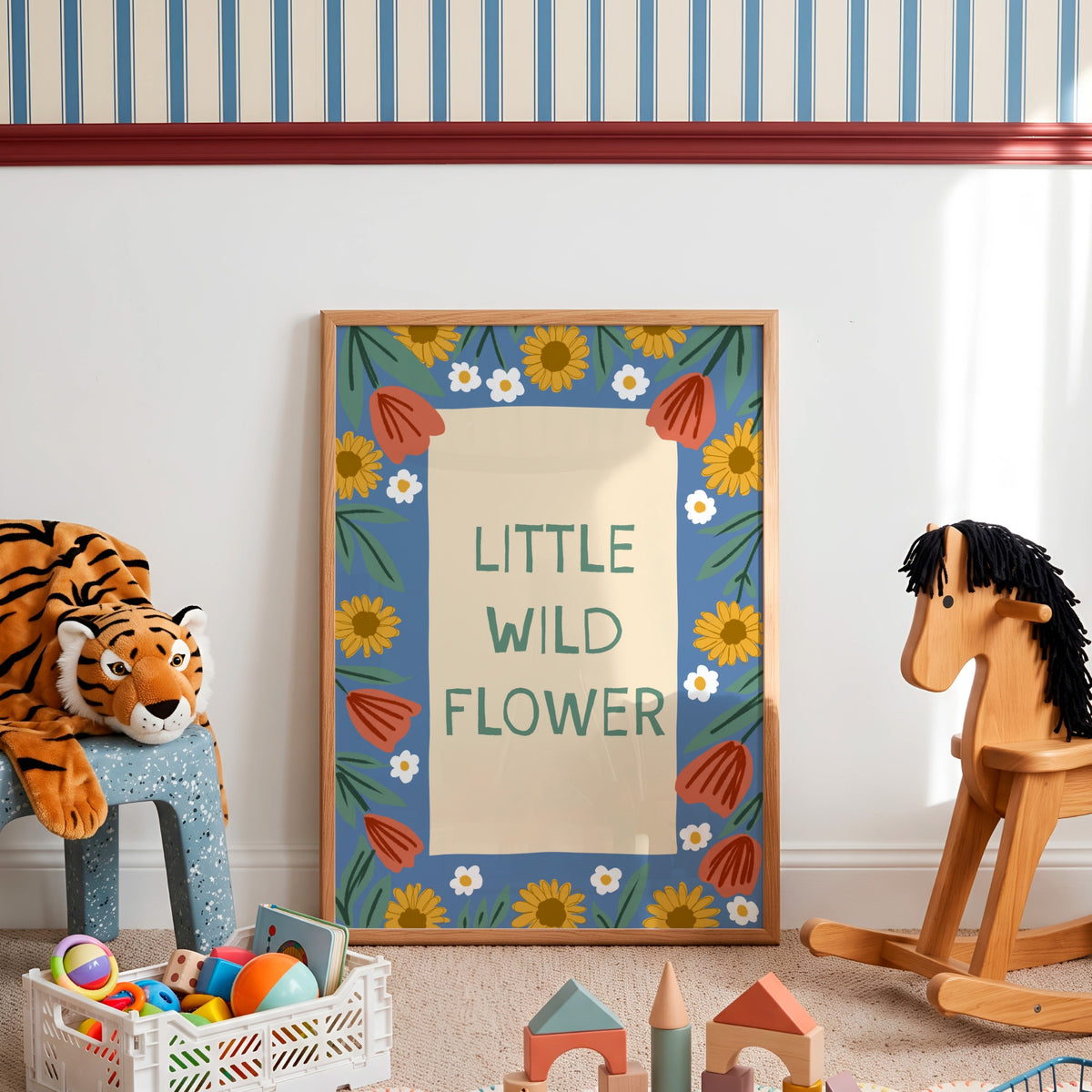 A framed children's art print reading 'LITTLE WILD FLOWER' with a blue floral border, displayed in a styled nursery or playroom setting with a tiger plush, wooden rocking horse, and toys around it.