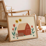 A framed art print featuring a whimsical barn illustration with yellow sunflowers and red tulips, styled in a warm, rustic children's room next to a wooden chair.