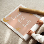 A person unrolling a large wall poster on a beige carpet. The poster has a dusty pink background with white text reading 'Dream Big little' and gold star and moon illustrations.