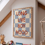 A framed wall sign with the phrase 'LONG LIVE BOY HOOD' in red letters over a blue and cream checkerboard pattern, displayed on a white wall in a child's room with toys and wooden beams visible.