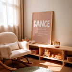 A framed nursery art print leaning on a light wood shelf in a sunlit nursery. The print has a dusty pink background with the word 'DANCE' in large white letters and smaller white text below.