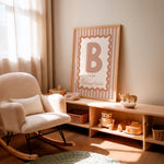 A framed nursery wall print with a large letter B and the phrase 'is for Ballerina' in blush pink and beige tones, leaning on a light wood shelf in a softly lit nursery next to a cream boucle rocking chair.