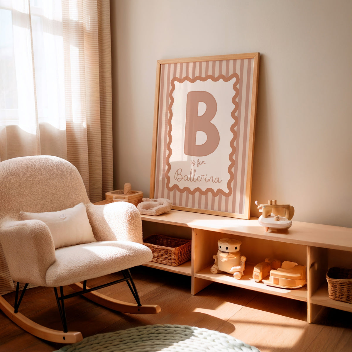 A framed nursery wall print with a large letter B and the phrase 'is for Ballerina' in blush pink and beige tones, leaning on a light wood shelf in a softly lit nursery next to a cream boucle rocking chair.