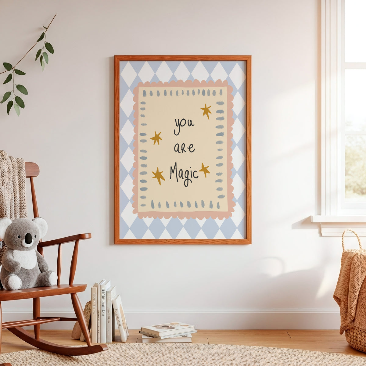 A framed nursery wall art print hanging on a white wall in a softly styled child's room. The print features the phrase 'you are Magic' in black script on a warm cream background, surrounded by a peach scalloped border and a light blue and white argyle pattern.