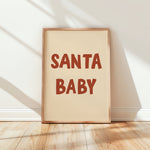 A framed art print with the words 'SANTA BABY' in a warm reddish-brown retro font on a cream background, leaning against a white wall on a light wood floor with soft window shadows.