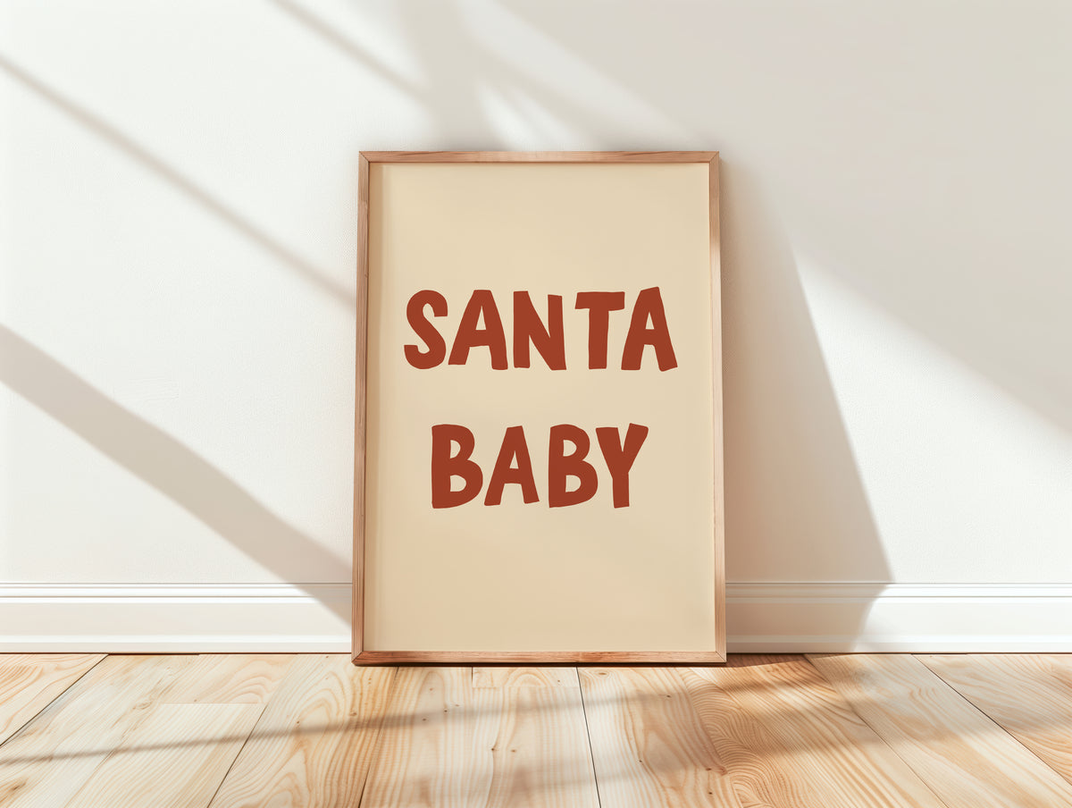 A framed art print with the words 'SANTA BABY' in a warm reddish-brown retro font on a cream background, leaning against a white wall on a light wood floor with soft window shadows.