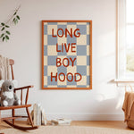 Framed wall art with the phrase “LONG LIVE BOY HOOD” in red serif letters over a blue and beige checkerboard pattern, displayed in a styled nursery room next to a wooden rocking chair and window.