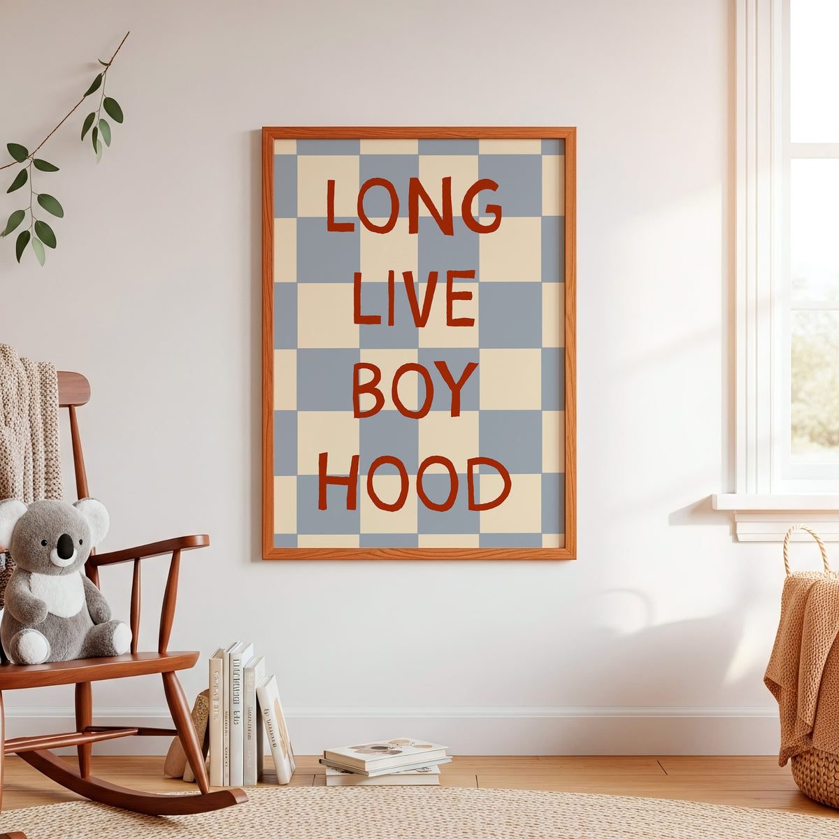 Framed wall art with the phrase “LONG LIVE BOY HOOD” in red serif letters over a blue and beige checkerboard pattern, displayed in a styled nursery room next to a wooden rocking chair and window.