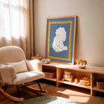 A framed art print featuring a white fluffy dog on a blue and yellow background, styled in a warm, sunlit nursery next to a cream-colored rocking chair and a wooden toy shelf.