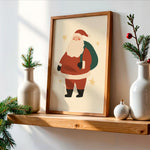 A framed art print of a cartoon Santa Claus hanging on a light-colored wall above a wooden shelf, styled with white ceramic vases, pine branches, and a small ornament.