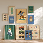 A gallery wall display of nine framed children's art prints with various colorful illustrations including trucks, a tiger on a skateboard, a bird, and motivational text, arranged on a light wood floor against a beige wall.
