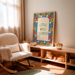 A framed nursery art print with the text 'LITTLE WILD FLOWER' in green on a cream background, surrounded by a colorful floral border. The print is displayed on a light wood shelf in a warm, sunlit nursery next to a cream-colored rocking chair.