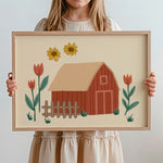 A child holds a framed art print featuring a whimsical barn illustration with flowers. The child is wearing a cream-colored dress, and the background is a clean, light gray studio wall.