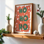 A framed vertical art print with the word 'HO HO HO' in green letters on a terracotta background with white polka dots, displayed on a wooden shelf in a styled home decor setting.