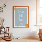 A framed nursery wall print with the words “Dream BIG little one” in white script on a light blue background, decorated with yellow stars and suns, hanging on a cream wall in a styled nursery room with a wooden rocking chair, books, and a plush toy.