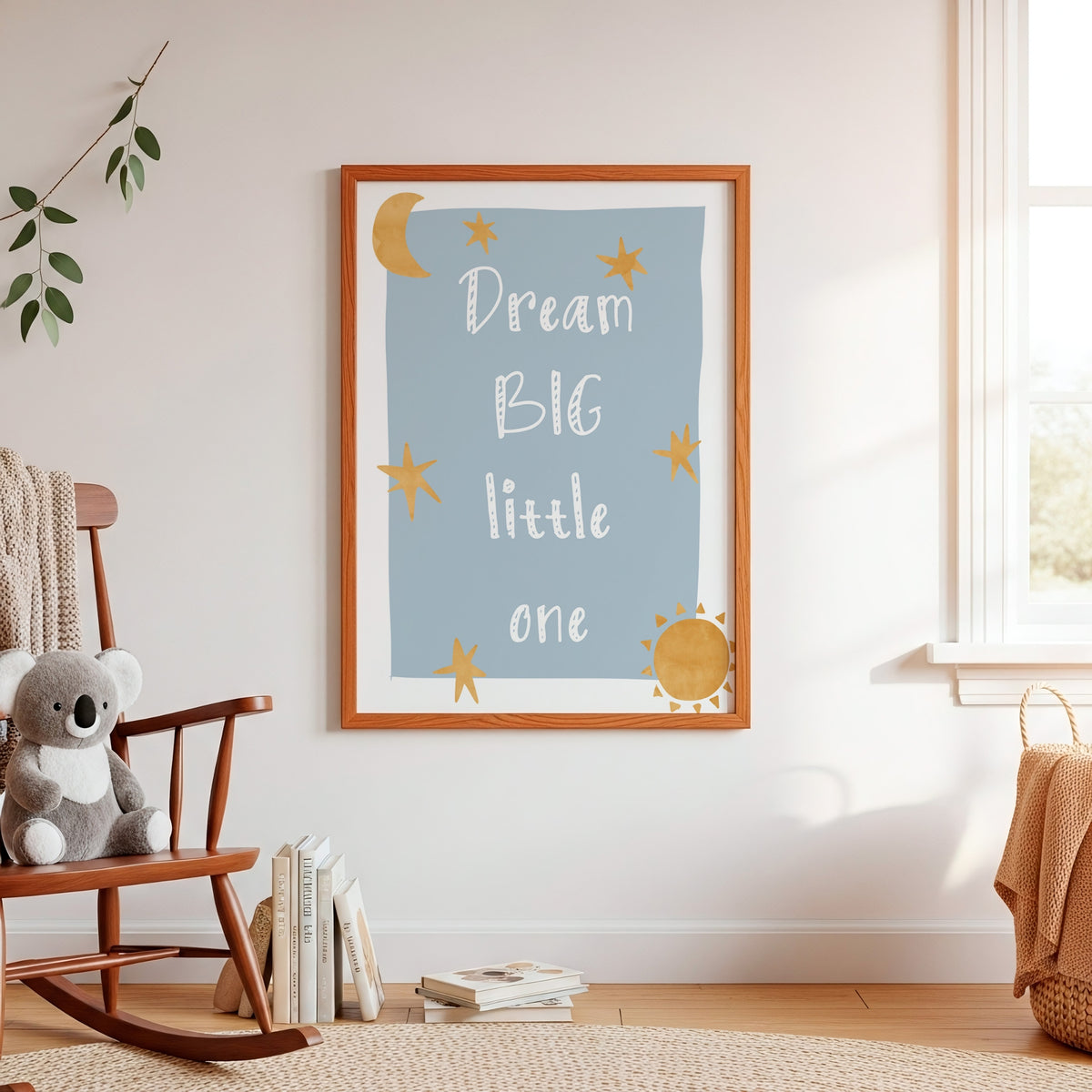 A framed nursery wall print with the words “Dream BIG little one” in white script on a light blue background, decorated with yellow stars and suns, hanging on a cream wall in a styled nursery room with a wooden rocking chair, books, and a plush toy.