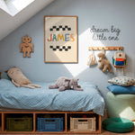 A styled children's bedroom corner with a low wooden bed, light blue bedding, plush toys, wall decor including a framed 'JAMES' print and 'dream big little one' sign, storage bins, pillows, and a stool.