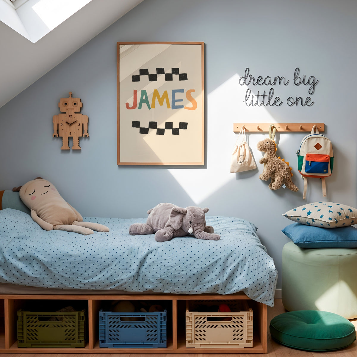 A styled children's bedroom corner with a low wooden bed, light blue bedding, plush toys, wall decor including a framed 'JAMES' print and 'dream big little one' sign, storage bins, pillows, and a stool.