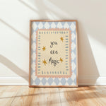 A framed art print leaning against a white wall on a light wood floor, featuring the text 'you are Magic' in black script with small gold stars, surrounded by scalloped peach and blue geometric borders.
