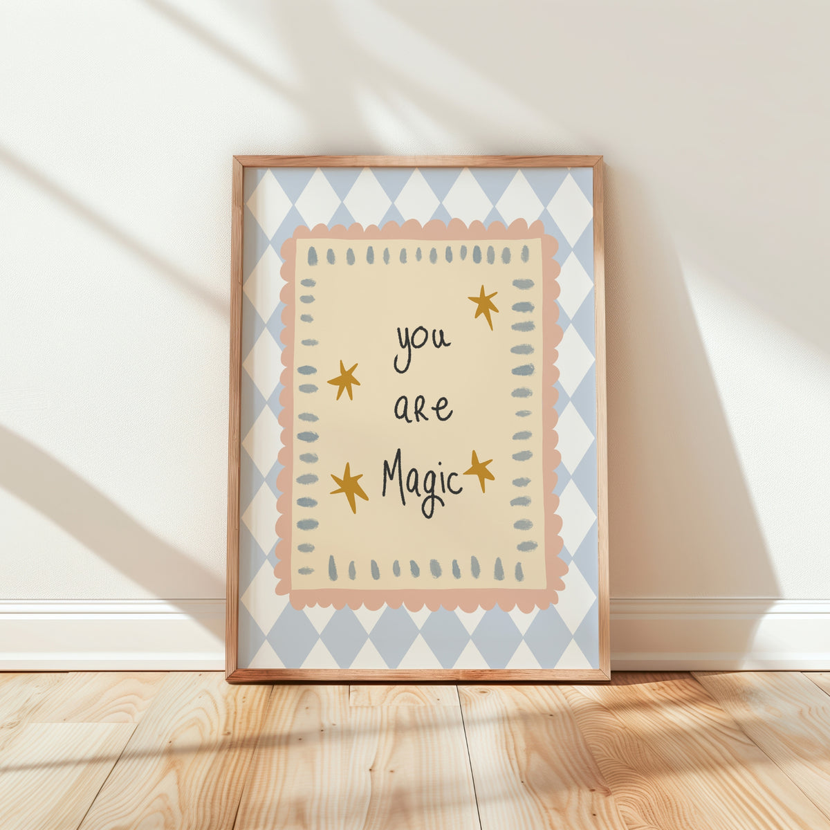 A framed art print leaning against a white wall on a light wood floor, featuring the text 'you are Magic' in black script with small gold stars, surrounded by scalloped peach and blue geometric borders.
