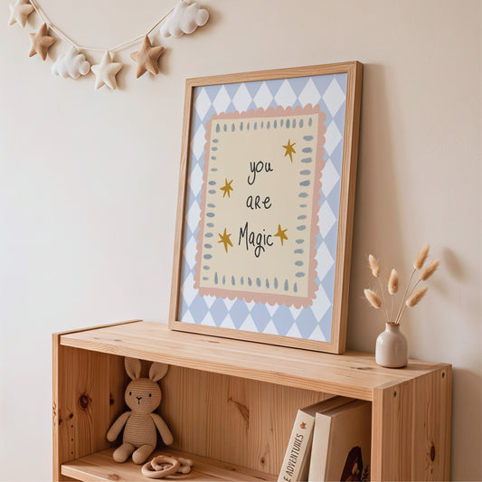 A framed art print with the text 'you are Magic' in black script, surrounded by yellow stars, displayed on a wooden shelf in a nursery setting.