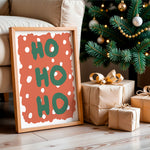 A framed Christmas art print with the words 'HO HO HO' in green on a terracotta background with white polka dots, leaning against a beige sofa next to a decorated Christmas tree and wrapped gift boxes.