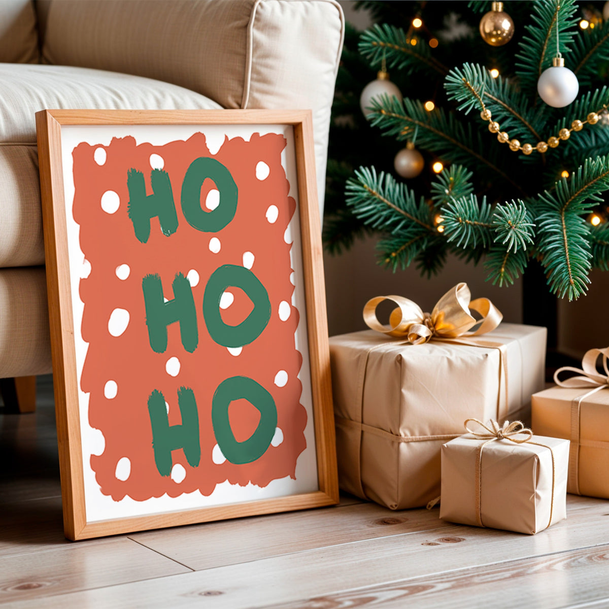 A framed Christmas art print with the words 'HO HO HO' in green on a terracotta background with white polka dots, leaning against a beige sofa next to a decorated Christmas tree and wrapped gift boxes.