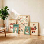 A set of five framed Christmas-themed art prints leaning against a beige wall in a sunlit room. The prints feature festive illustrations including Santa Claus, Rudolph the Red-Nosed Reindeer, a Christmas tree, an alphabet board with holiday icons, and a sign reading 'SANTA BAE'.