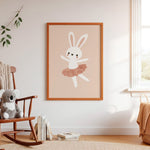 A framed nursery art print featuring a white bunny in a dusty pink tutu on a warm beige background, hanging on a white wall in a styled nursery room with a wooden rocking chair, plush toy, and window.