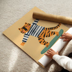 A person's hands are unrolling a large poster or wall art print on a beige carpet. The artwork features a cartoon tiger wearing a black and white striped shirt and riding a green skateboard, with a brown cardboard mailing tube resting on top of the print.